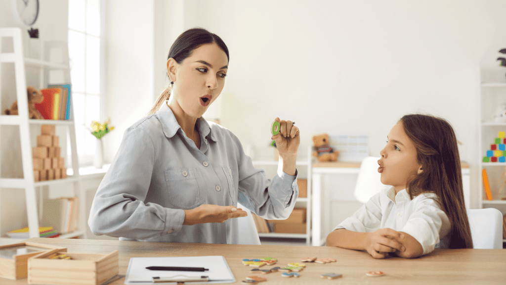 A teacher stands in front of a classroom, pointing at a large phonetics chart. The chart illustrates various letters and their corresponding phonetic sounds. The teacher explains the sounds to a group of attentive students seated at their desks, who follow along with their own mini phonetics charts.